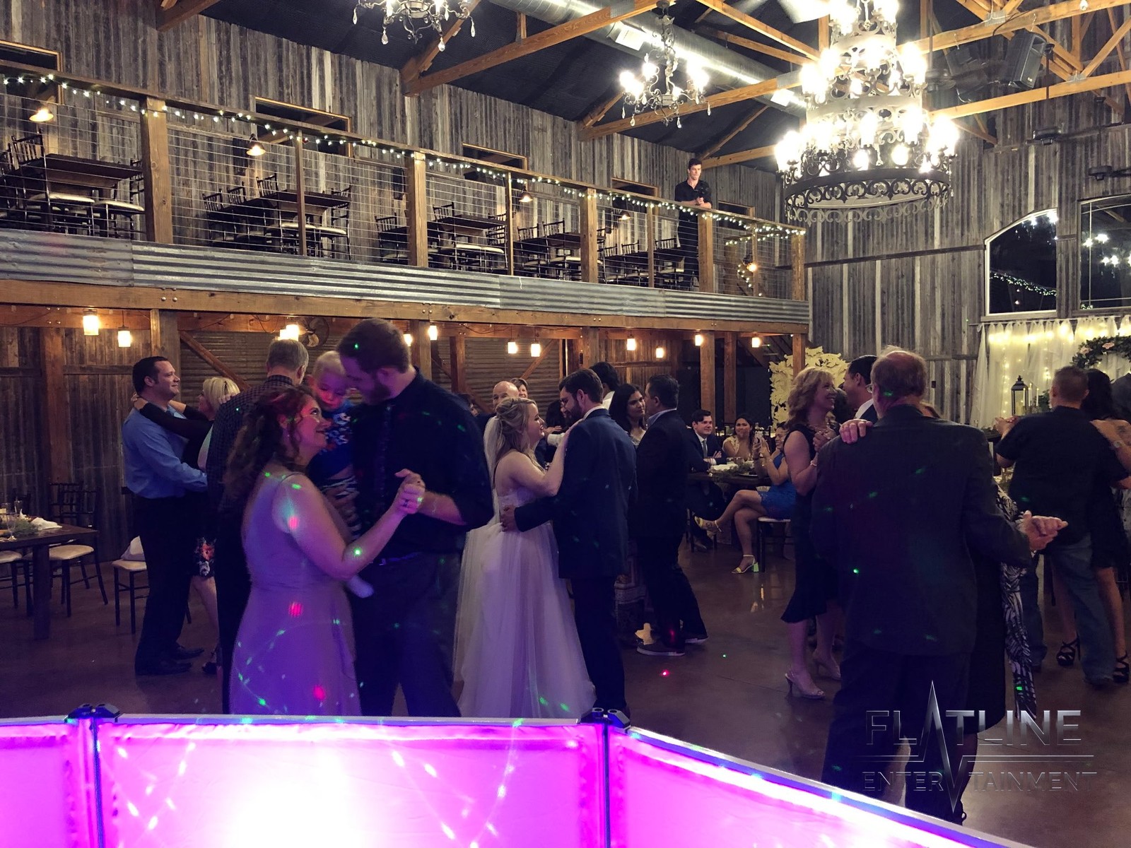 A wedding reception dance floor in a rustic barn venue with the DJ booth glowing pink in the foreground